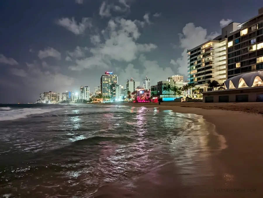 A crescent-shaped beach at night with lit-up buildings in the background.