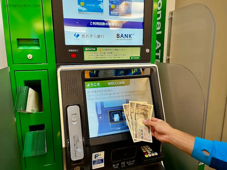 A hand holding a couple of Japanese yen bills next to an ATM.