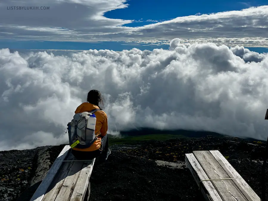 A hiker sitting town and looking at a view with clouds beneath her.