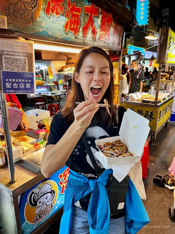 A woman standing next to a food stalls with an octopus leg in between a pair of chopsticks.