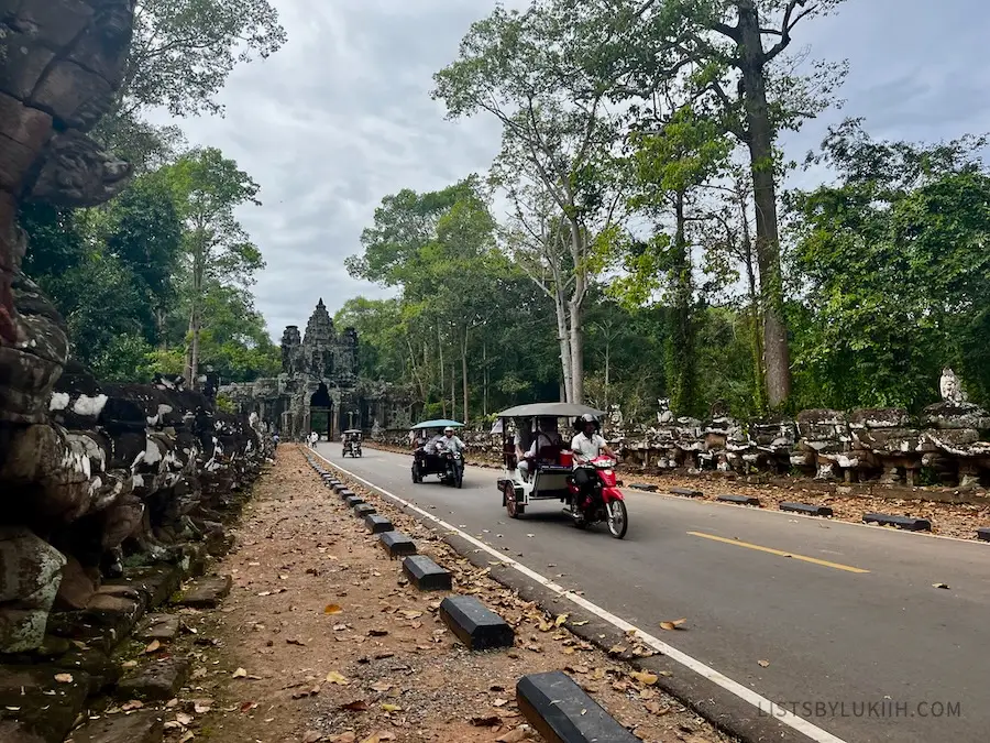 A few tuk-tuks going down a two-lane road with ancient temples behind them.
