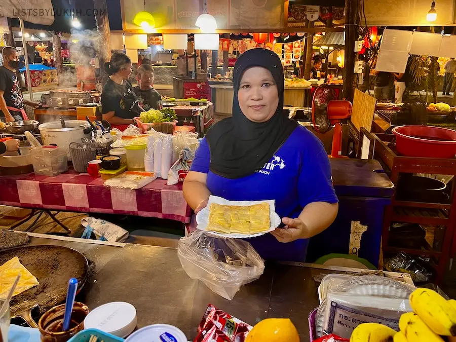 A woman holding a flat pancake in an outdoor market with many food stalls.