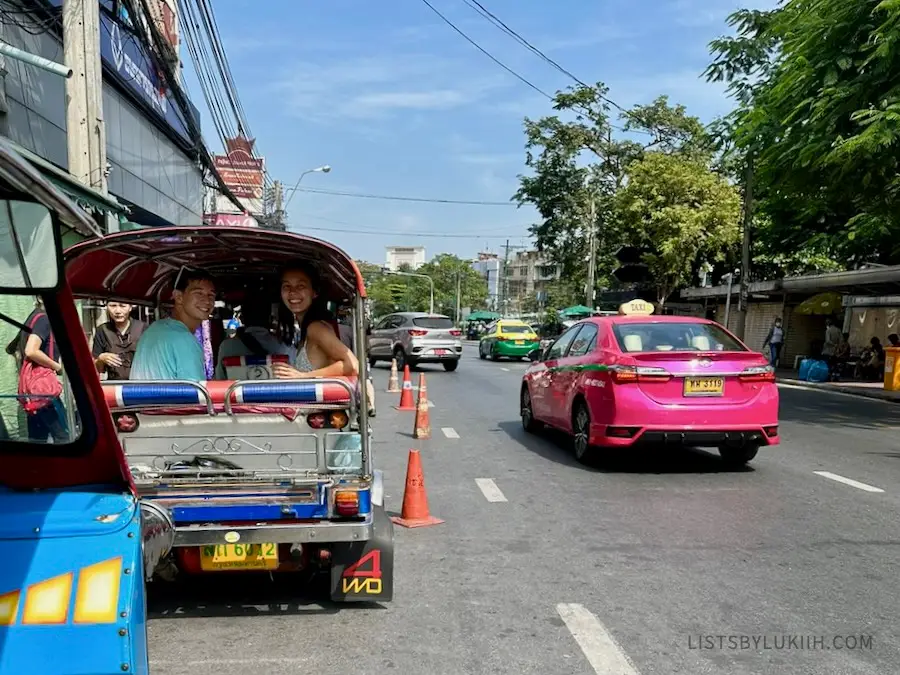 A street with cars and people riding tuk-tuks.