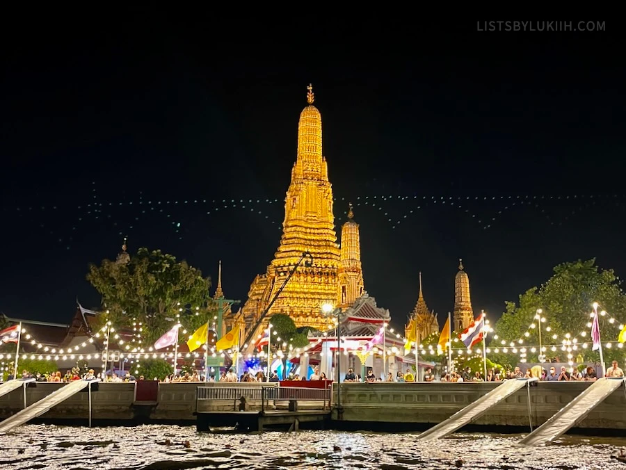 A gold temple lit up at night overlooking a river with many people surrounding the area.