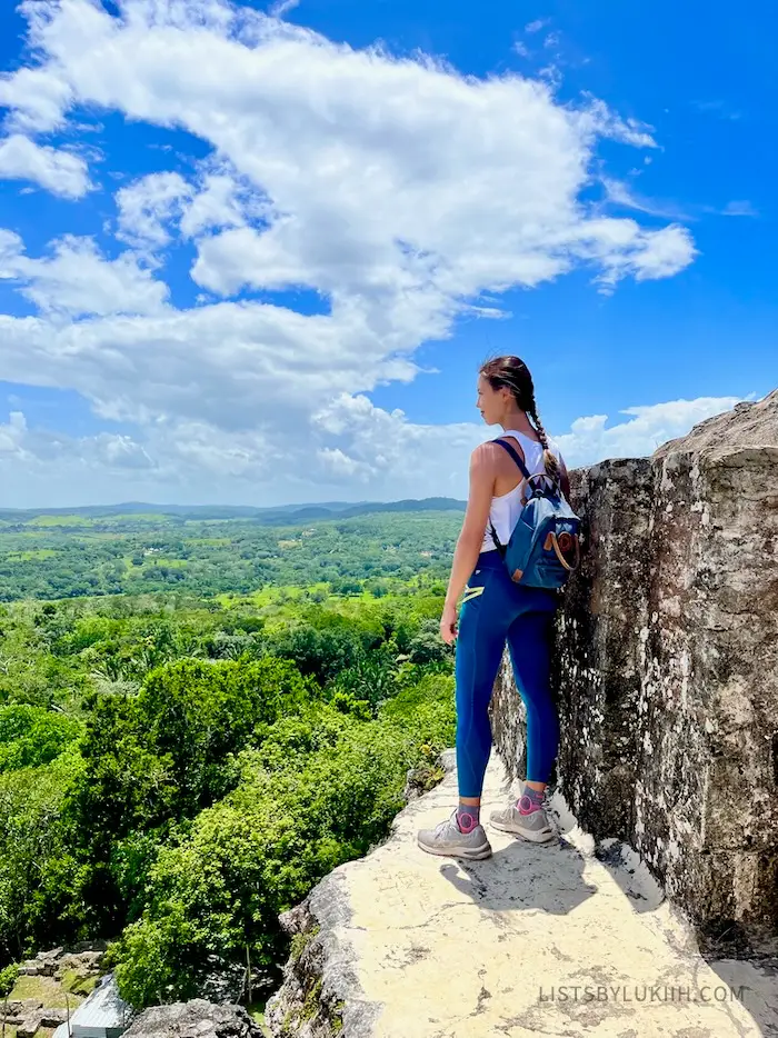 A woman standing on a ruin's cliff overlooking lush trees.