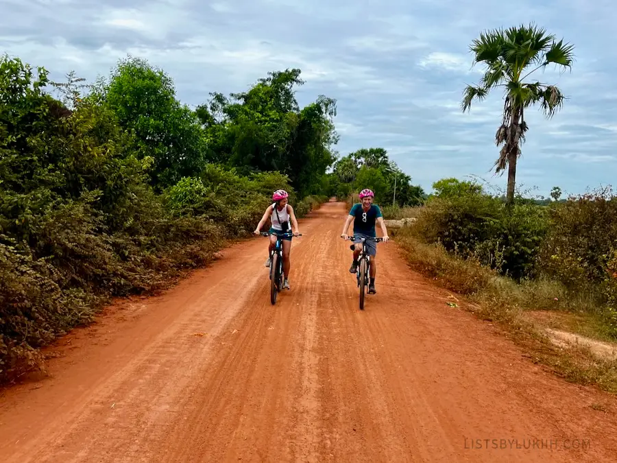Two people riding bikes down a red dirt path surrounded by bushes.