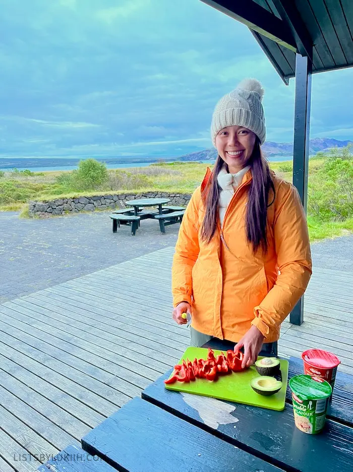 A woman cutting bell peppers on a cutting board outside with nature and mountains in the backround.