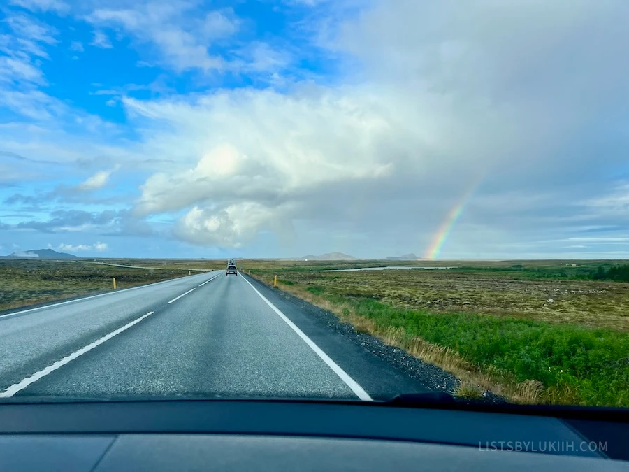 A road from the perspective of a vehicle winding through grass field with a rainbow on the horizon.