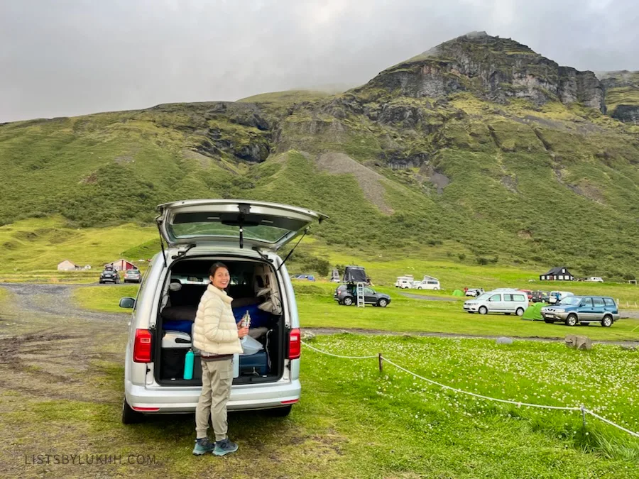 A woman standing in front of a campervan parked next to a mountain with other campervans in the distance.
