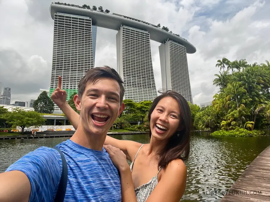 Two people taking a selfie in front of a tall building with three structures with a connected ceiling.