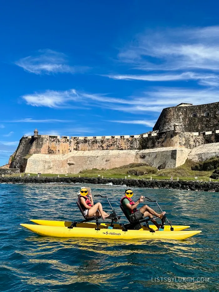 Two people sitting on a water bike in an ocean with a fort in the background.