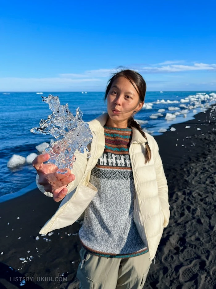A woman holding a crystal-like ice chunk with black sand beach in the background.