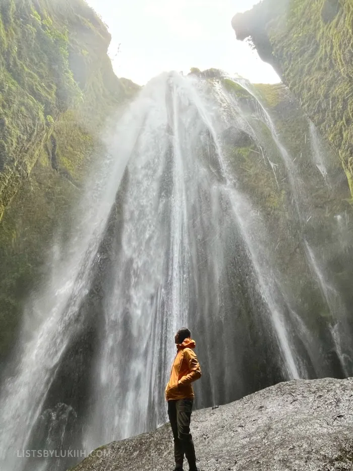 A woman standing inside a cave looking up at a tall waterfall.