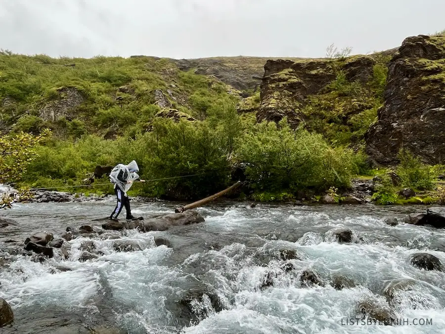 A man wearing a poncho crossing rocks and a log in a moving river.
