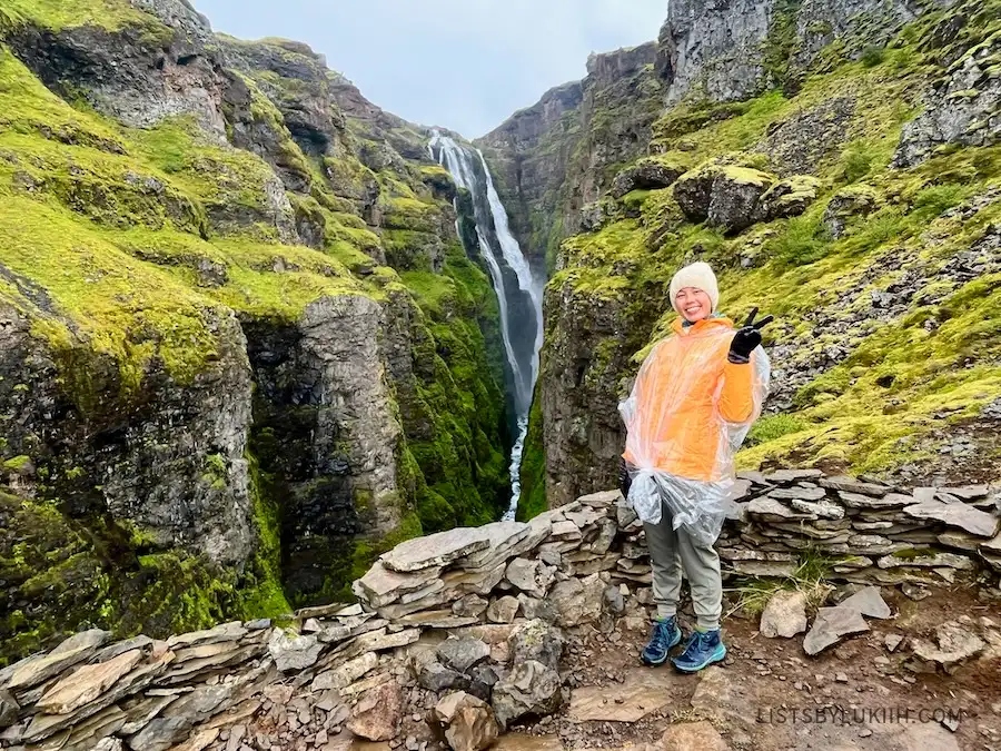 A woman wearing a poncho and a raincoat in front of a waterfall in between cliffs.