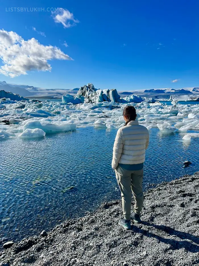 A woman staring at a lagoon with giant icebergs floating in it below a blue sky.