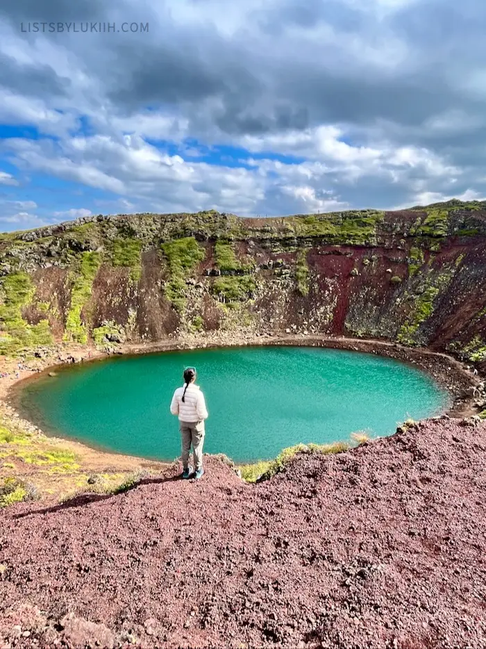 A woman standing near the edge of a crater with blue water filled at the bottom.