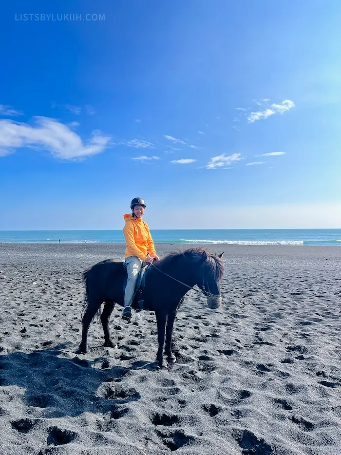 A woman riding a small horse on a black-sand beach.