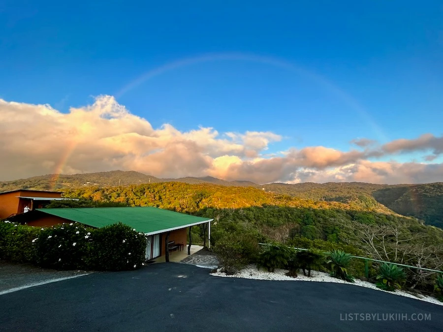 A giant rainbow against a sky over a valley with a cabin hotel on the left side.