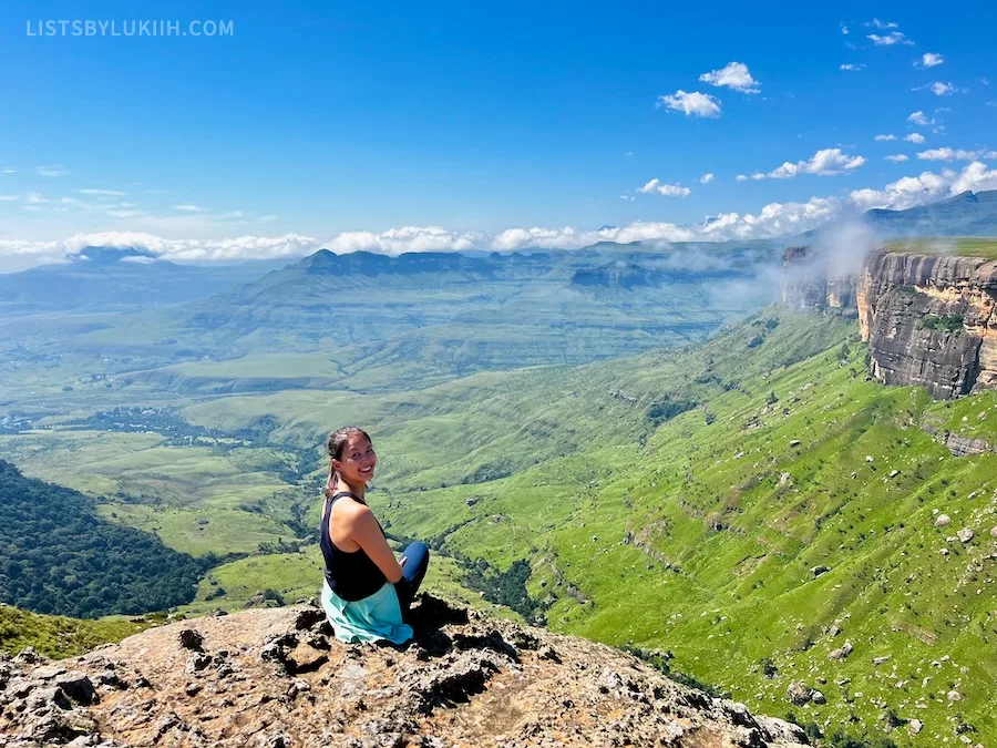 A woman sitting at the edge of a mountain cliff overlooking a lush valley with clouds in the horizon.