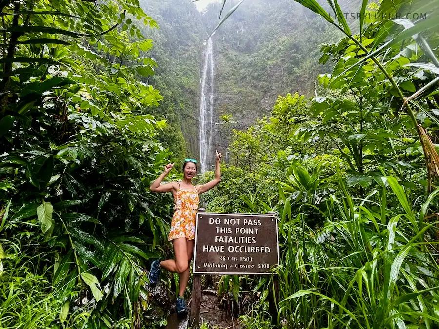 A woman standing next to a sign that says it's dangerous to approach the waterfall in the background.