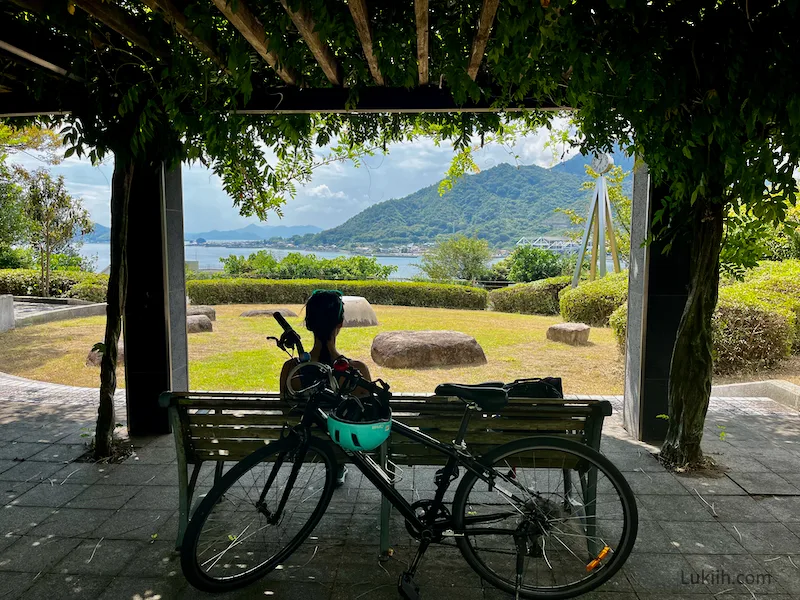 A woman sitting on a bench next to a bike looking out at a park and ocean view.