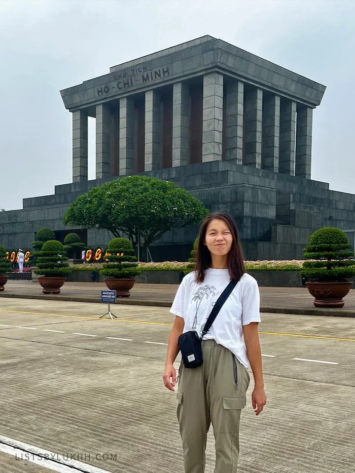 A woman standing in front a box-shaped, gray building with columns that says "Ho Chi Minh."