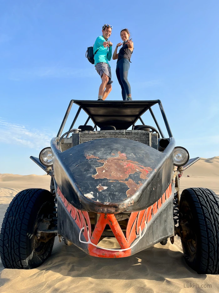 Two people standing on a roof of an open vehicle on sand.