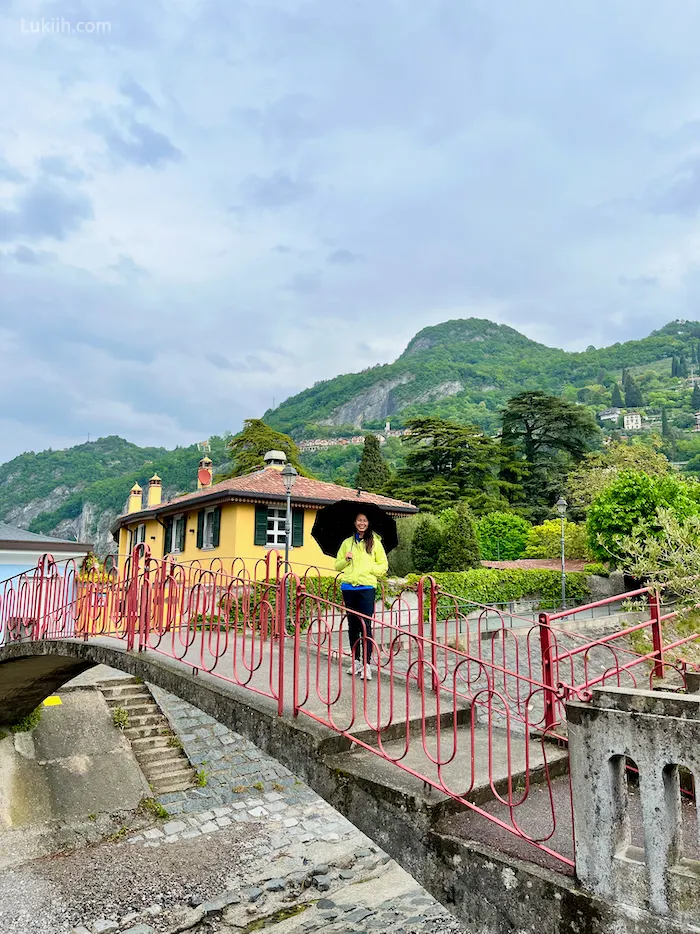 A woman standing on a red bridge with a lush, green mountain behind her.