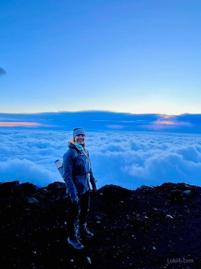 A woman standing on a mountain edge with clouds in the background.