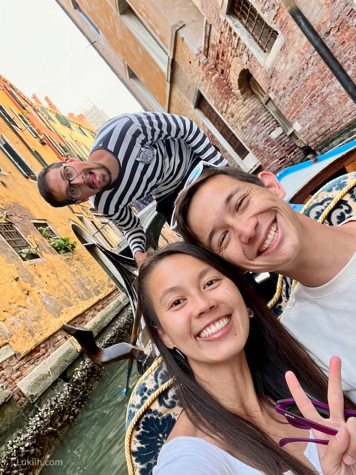 Three people taking a selfie on a boat in a small canal. One man is standing and wearing a striped shirt.