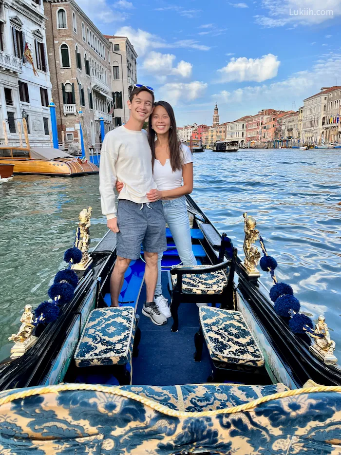Two people standing on a blue boat in a big canal surrounded by colorful buildings.