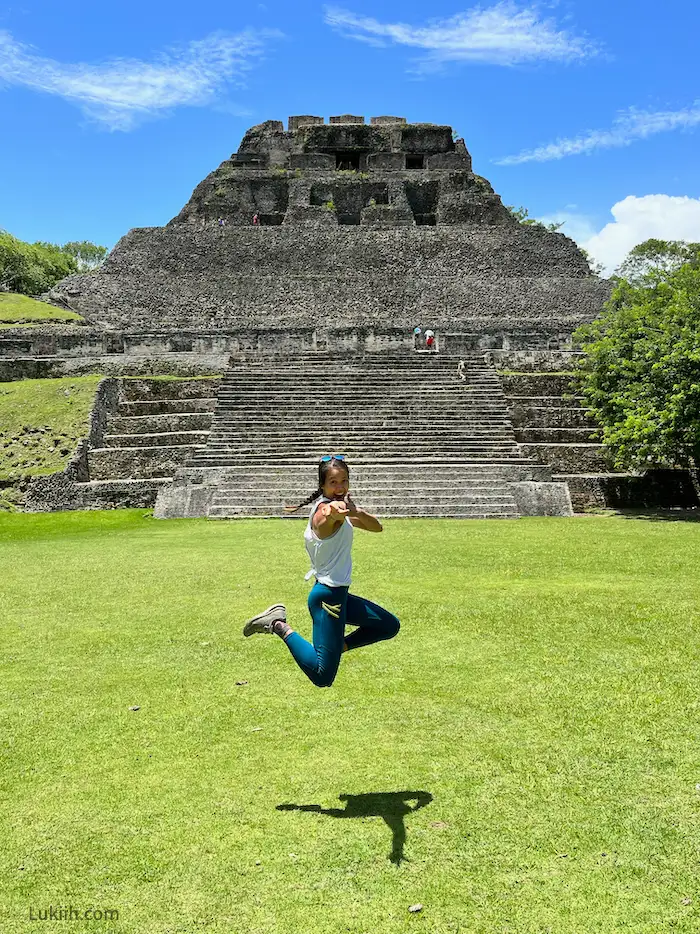 A woman jumping in front of a big Maya ruin made of stone with stairs.
