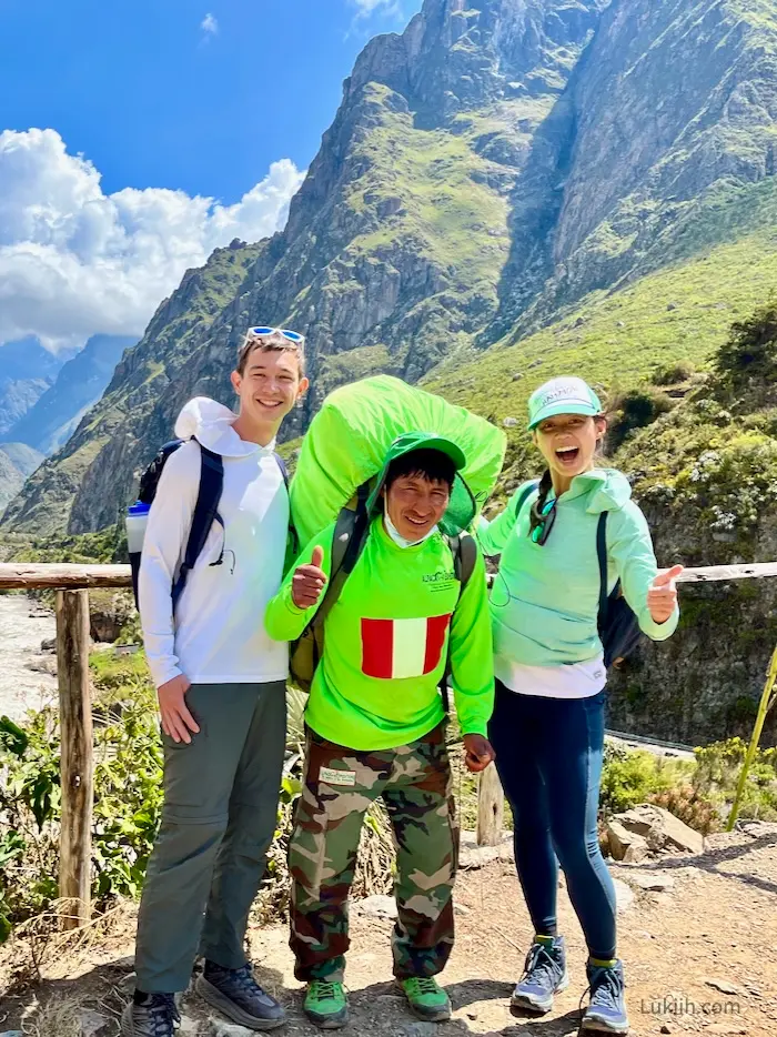 Two tourists on a mountain and a porter carrying a large bag is between them.