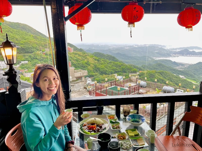 A woman holding a small cup of tea sitting next to a view of mountains and red lanterns.