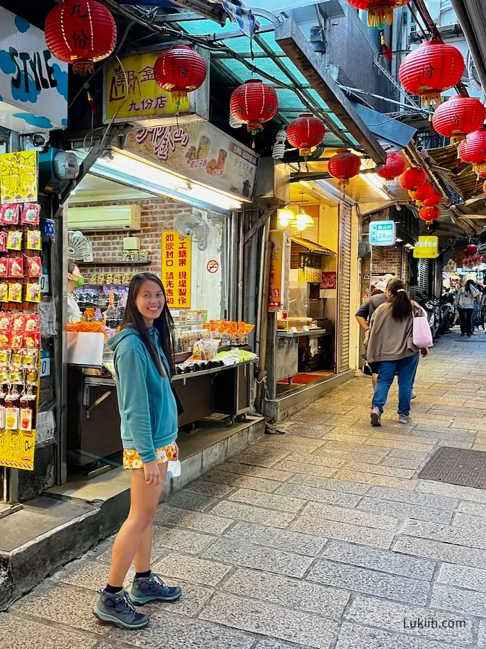 A woman smiling at the camera with red lanterns and small vendor shops in the background.
