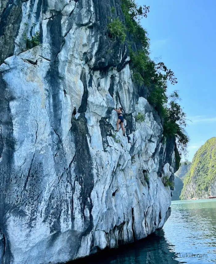 A woman climbing up a gray limestone rock over water.