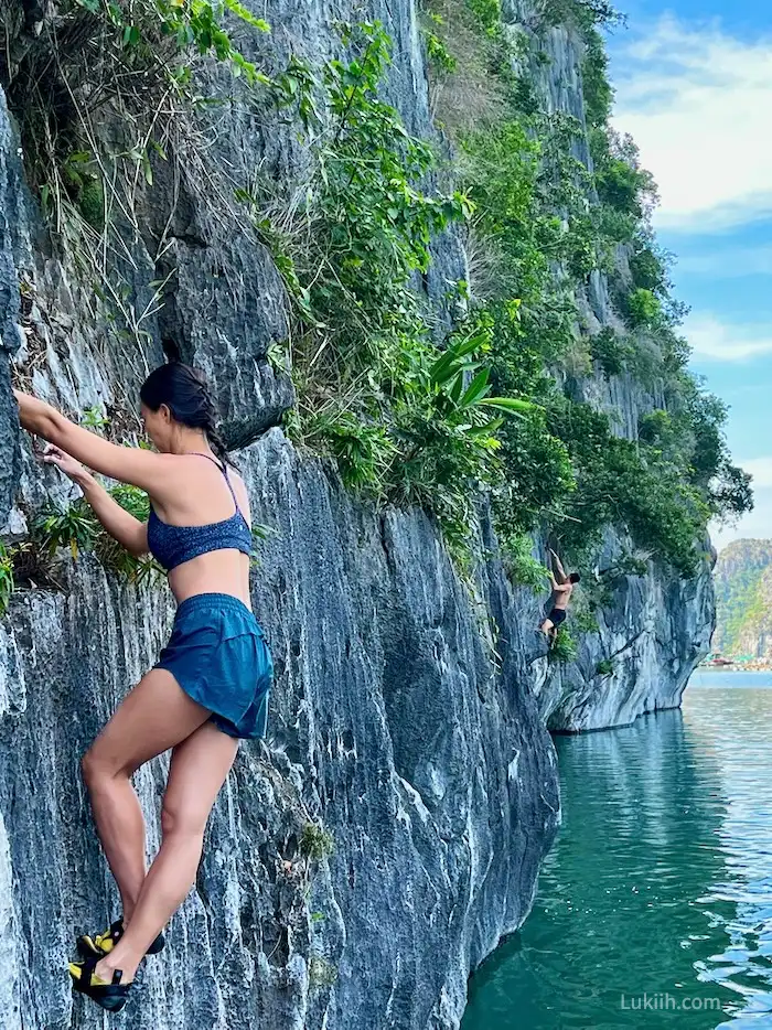 A closeup of a woman climbing up a gray limestone rock over water without any ropes.