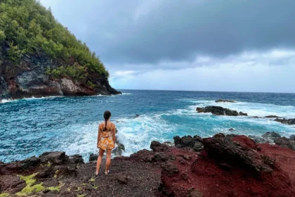 A woman standing on red sand with a very blue ocean in the background.