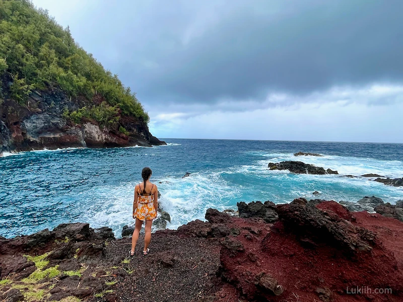 A woman standing on red sand with a very blue ocean in the background.