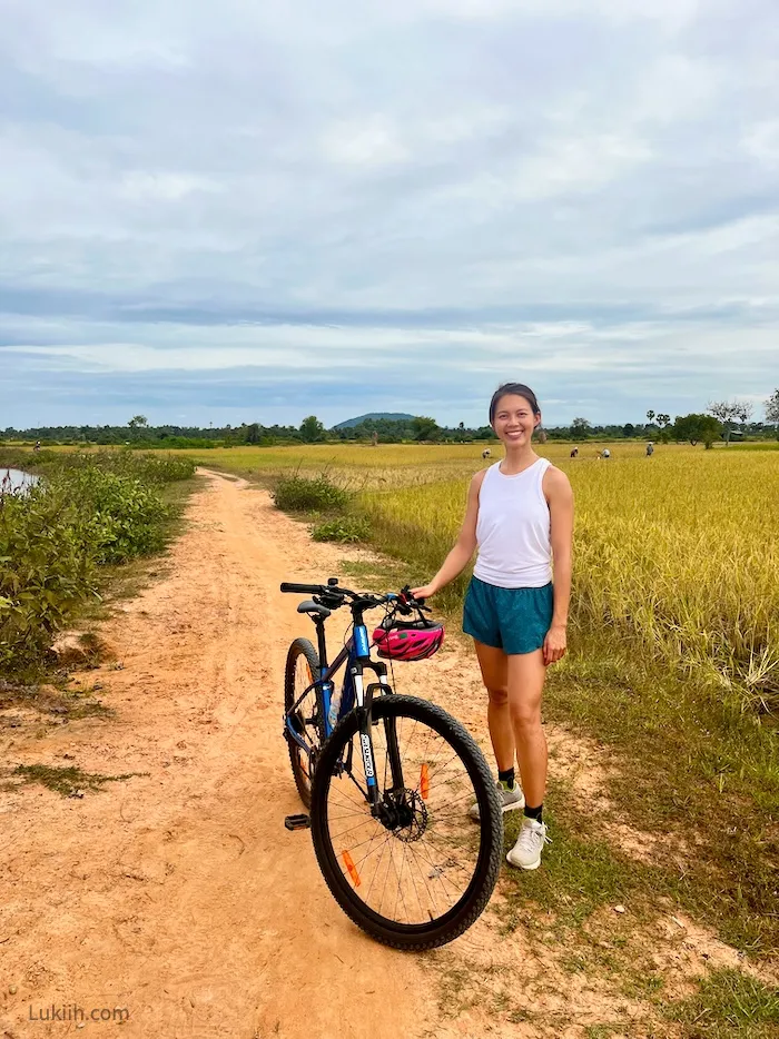 A woman standing next to a bike with rice paddy fields in the background.