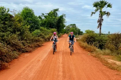Two people riding bikes down a red dirt path surrounded by trees and bushes.
