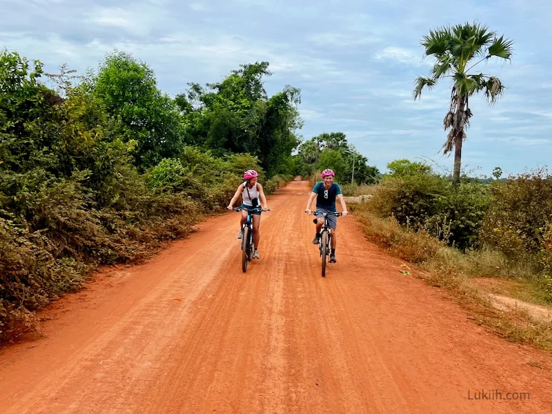 Two people riding bikes down a red dirt path surrounded by trees and bushes.