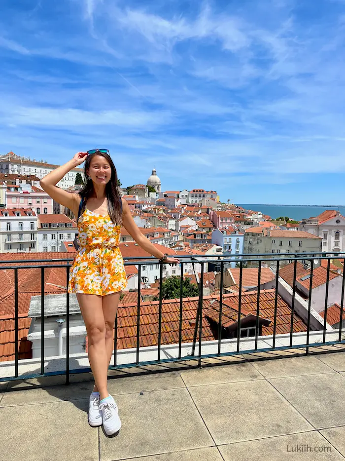 A woman on a balcony overlooking a background with buildings with orange-clay roofs.