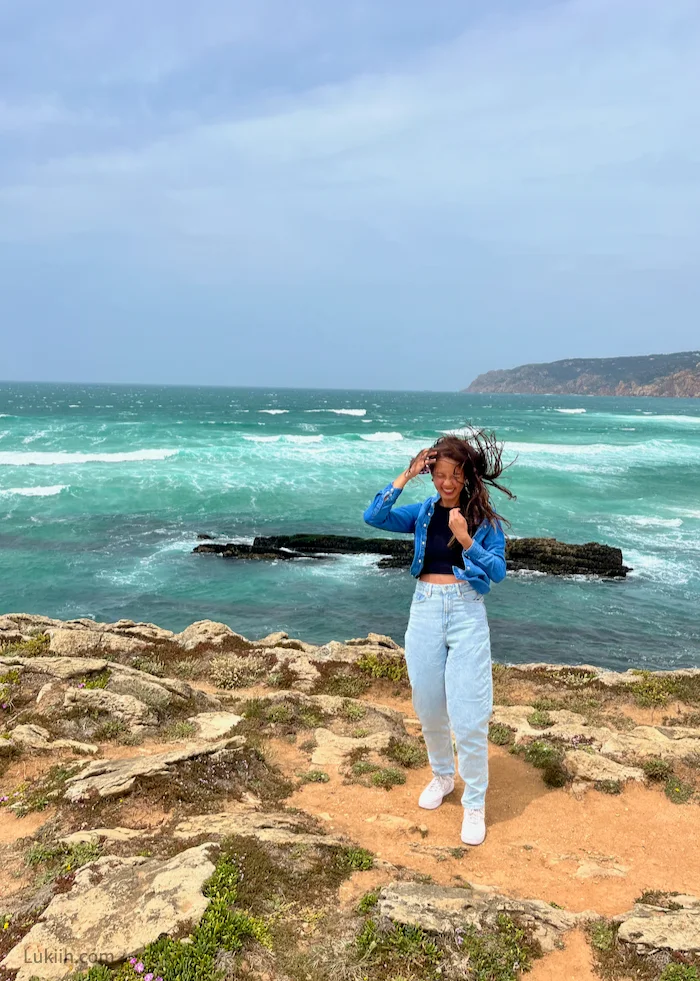 A woman standing on rocky sand by a teal ocean while the wind blows her hair.