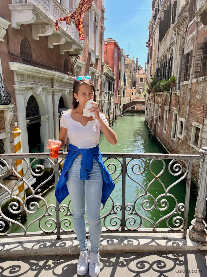 A woman taking a bite out of a crepe while standing in front of a narrow water canal.