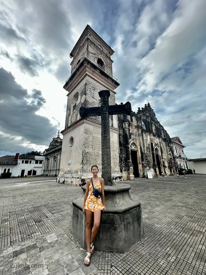 A woman standing next to an old cross with a church behind them.