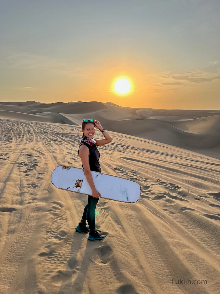 A woman standing in a desert holding a sand board.