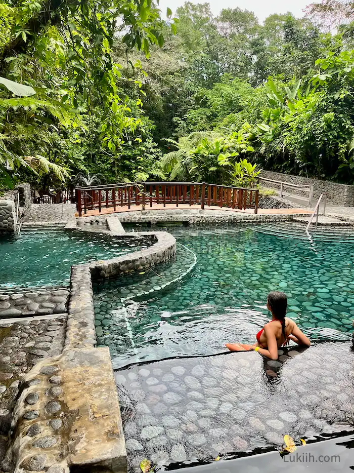 A woman leaning in a quiet pool with a bridge and lush trees in the background.