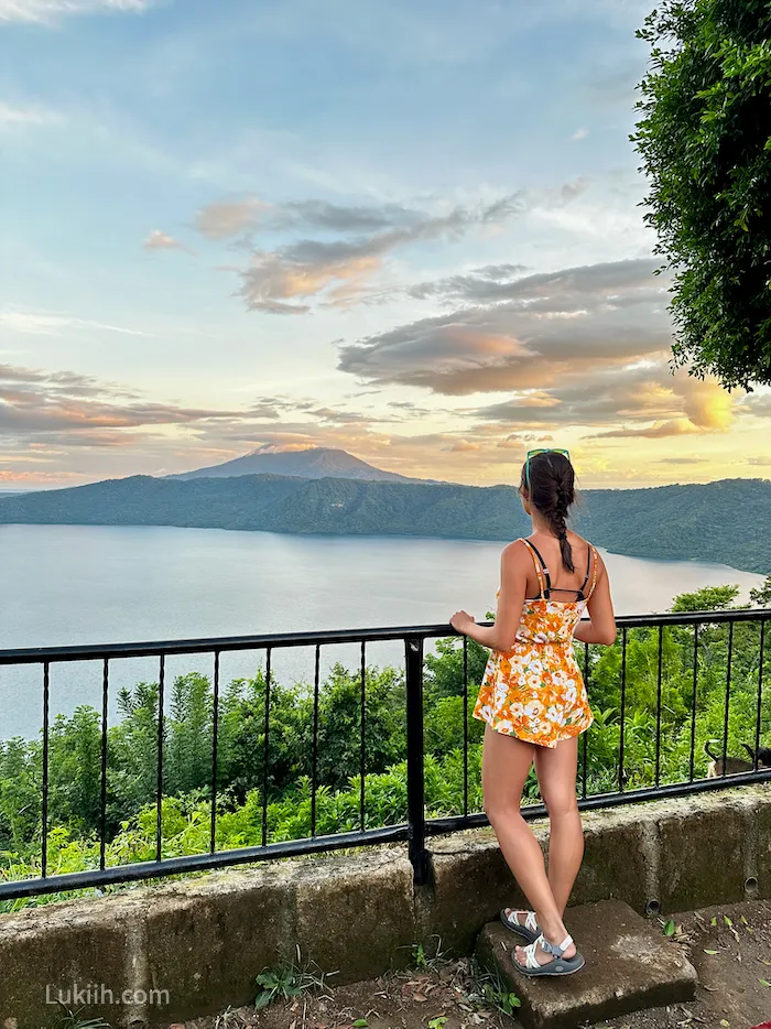 A woman standing at a viewpoint looking out at a volcano and clear lake.